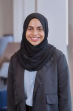 Mas in a blue shirt, black blazer, and black hijab. Smiling and standing in front of a blurred background.