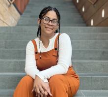Woman sitting on steps with braids and glasses; white long-sleeved shirt and orange/brown overalls