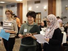 Photo of three students standing near the middle of a room, smiling, as others around the room talk and move about